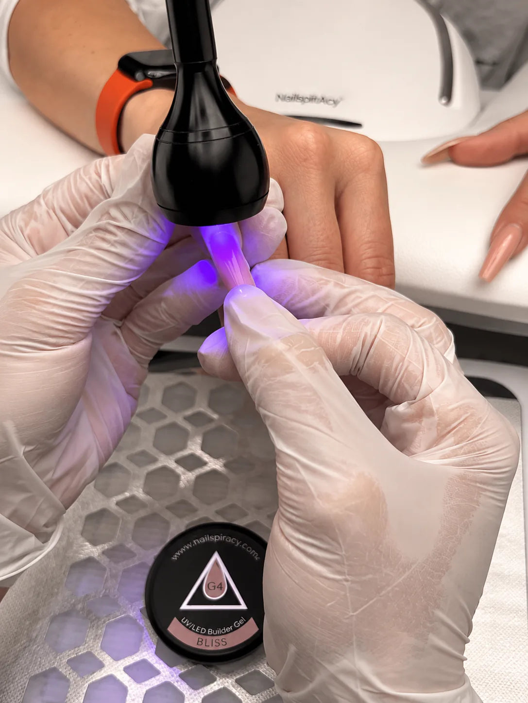 Gloved nail technician cures gel inside a dual nail form under a compact UV lamp during a professional manicure demonstration.