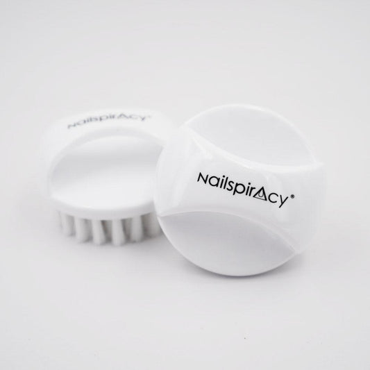 Two white, round-shaped nail dust brushes with bristles, one standing upright and the other lying on its side, with the brand name 'Nailspiracy' visible on the side of the standing brush.