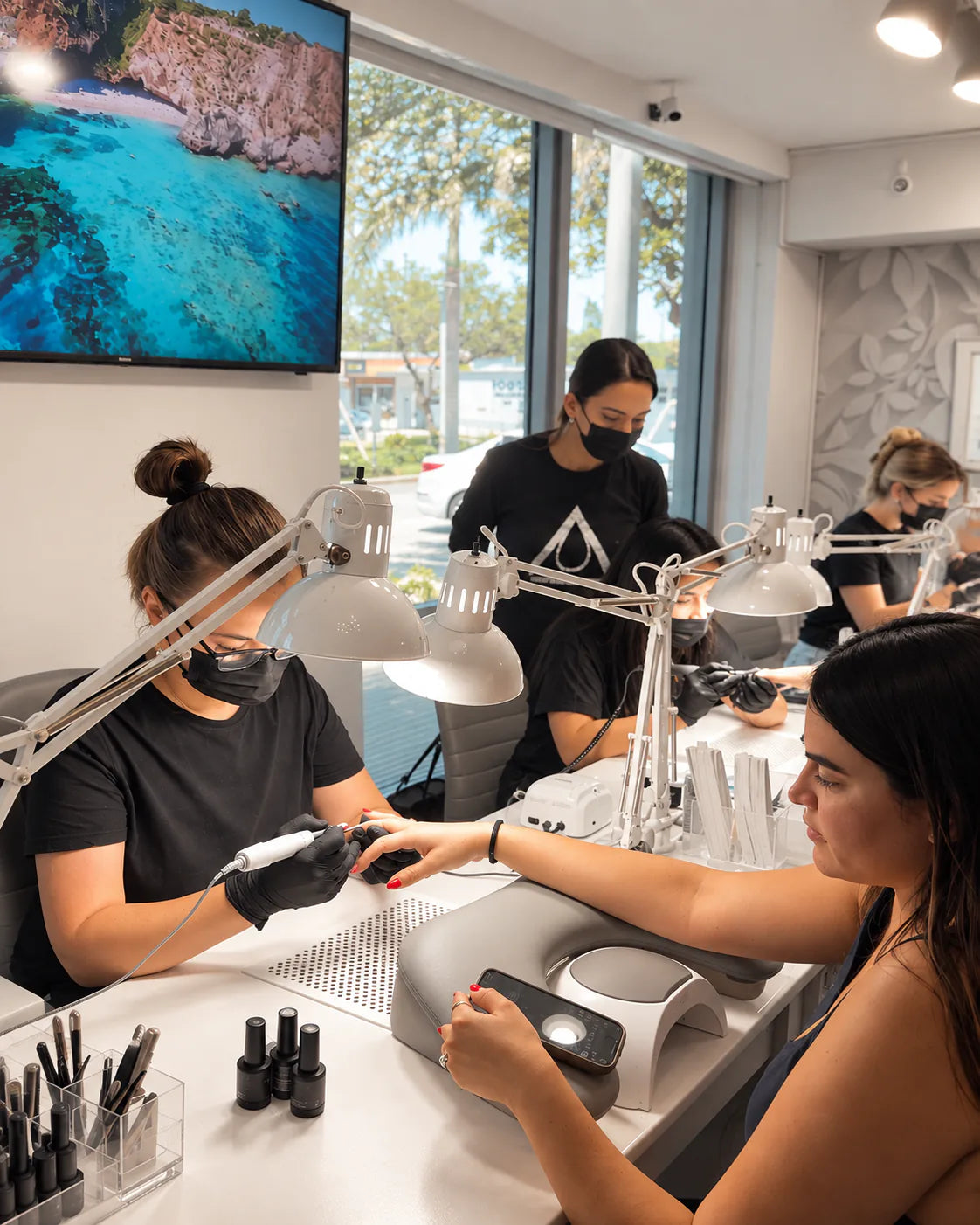 Bright nail salon during a Nailspiracy Russian Manicure From Zero class, with Siranush Ambartsumian teaching students at manicure stations while a student performs a manicure.